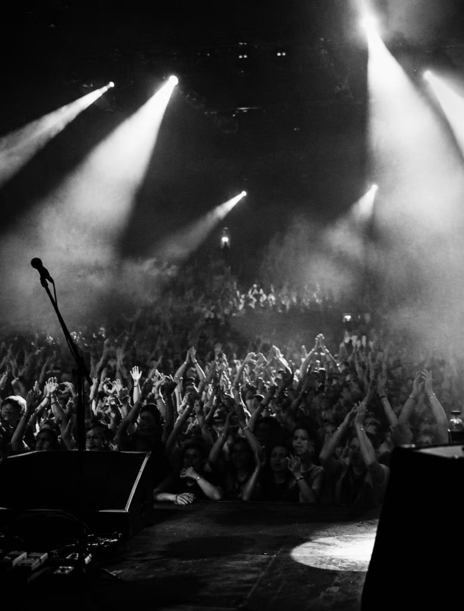 A concert crowd seen from the stage with beams of light cutting through haze