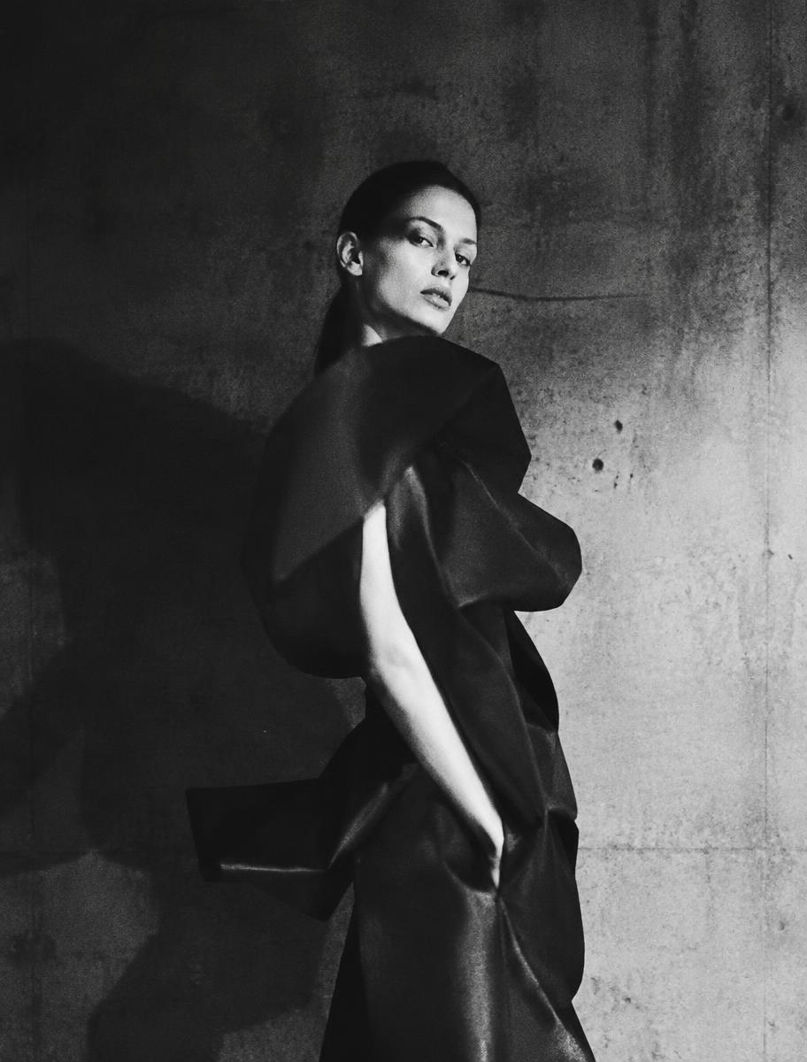A model in a sculptural dark garment against a raw concrete wall